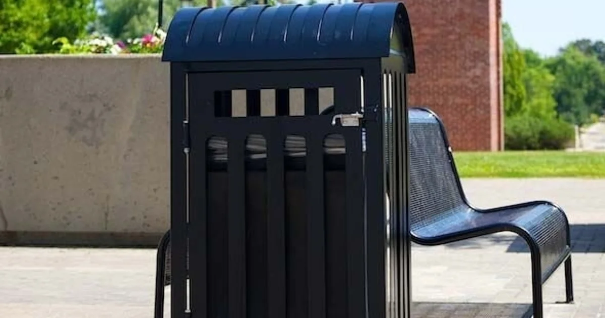 Black metal trash bin beside a curved bench in a park.